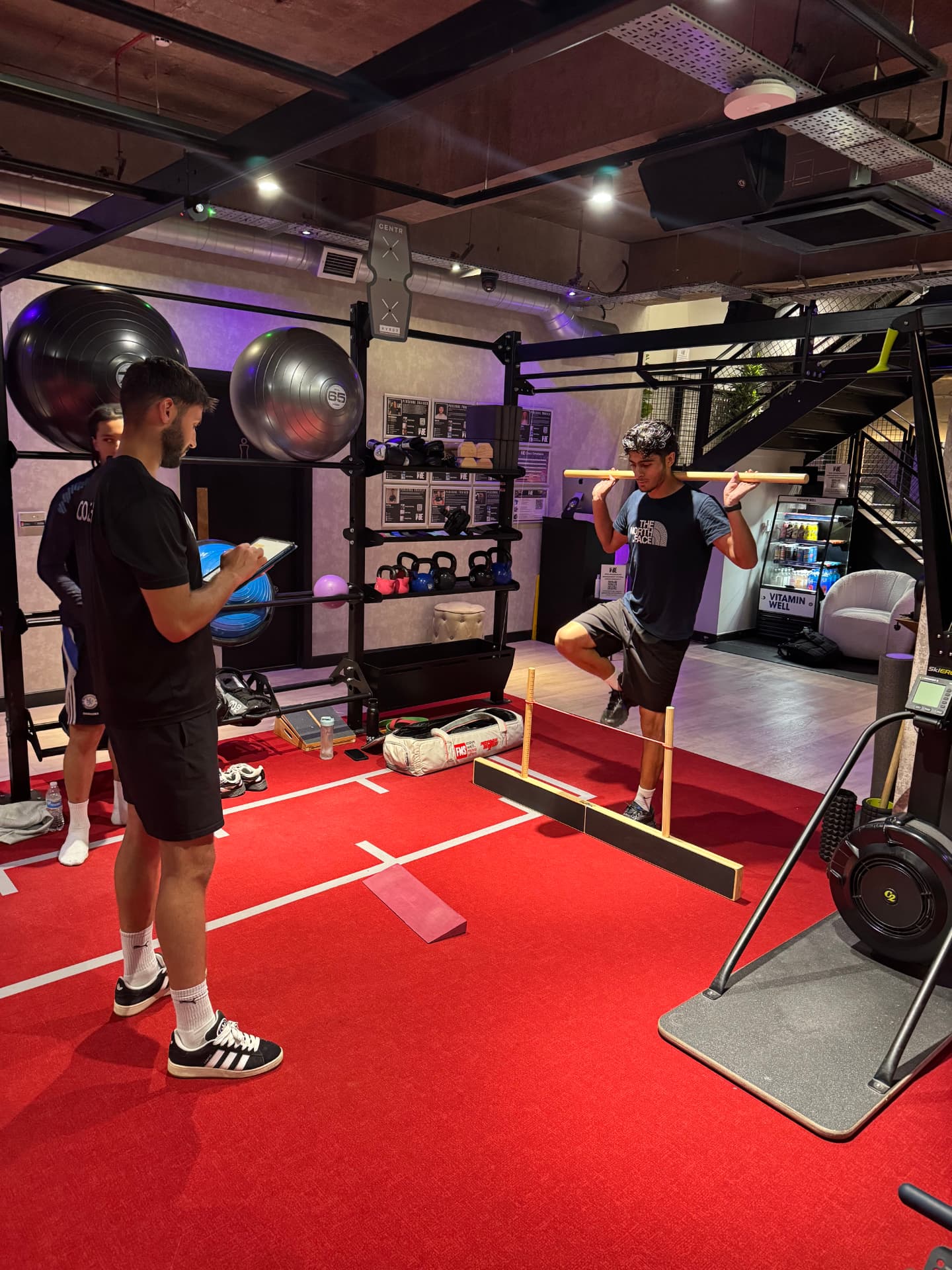 Coach watching a client step over a hurdle cord during a movement screening, clipboard in hand